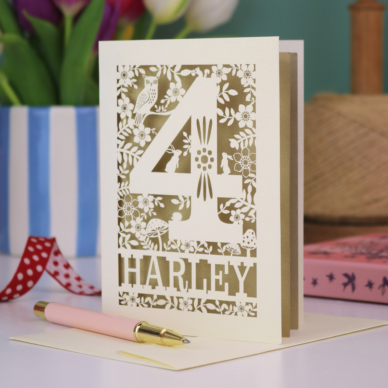 Decorative 4th Birthday card with floral and animal laser cut patterns, personalised with the name 'Harley', on a table with stationery items.