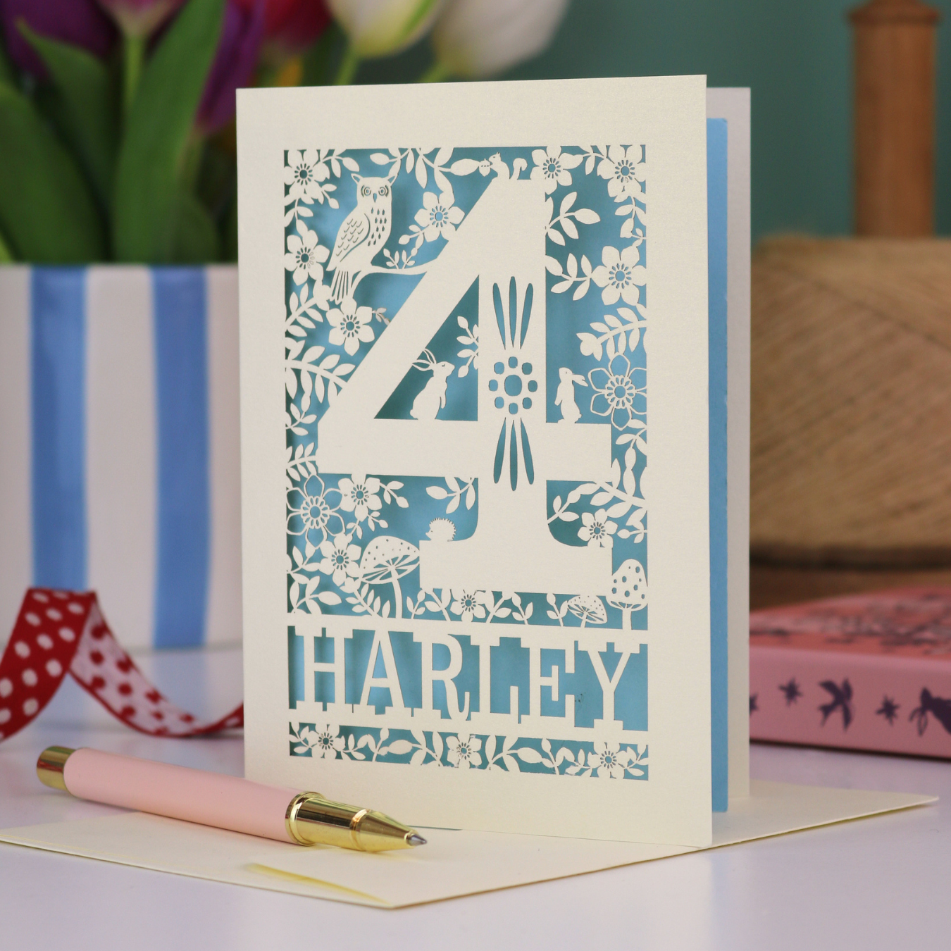 Decorative 4th Birthday card with floral and animal laser cut patterns, personalised with the name 'Harley', on a table with stationery items.