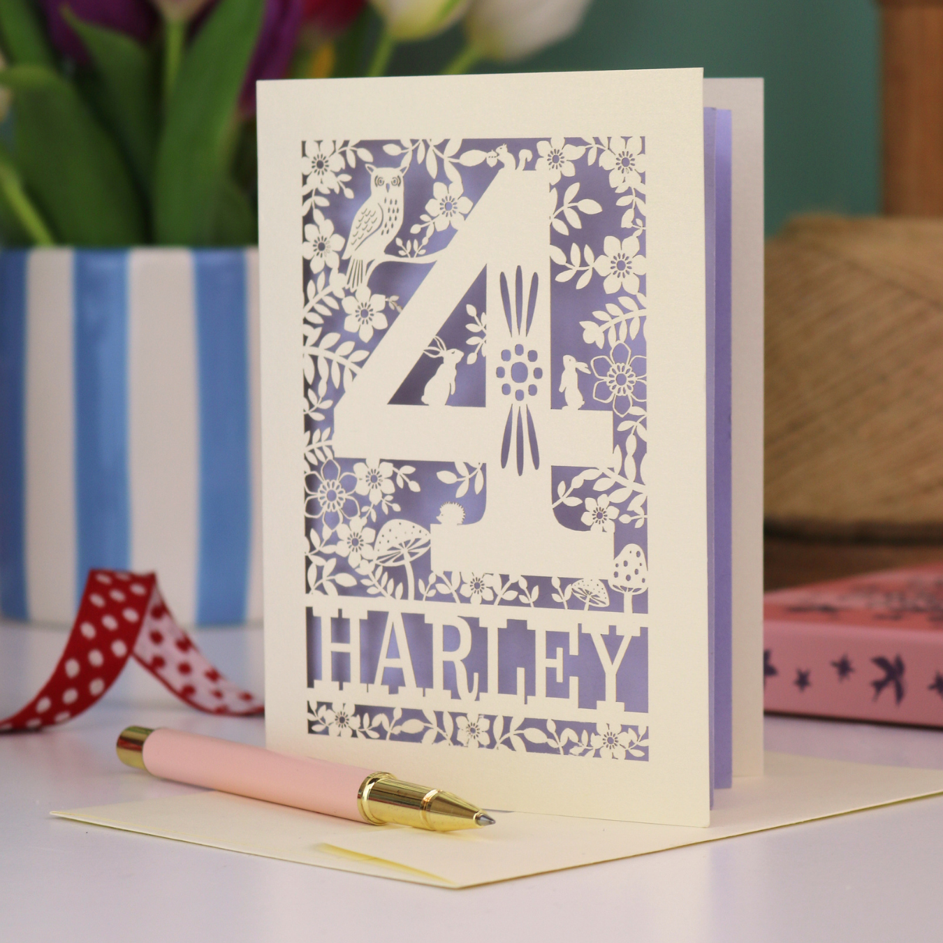 Decorative 4th Birthday card with woodland laser cut patterns, personalised with the name 'Harley', on a table with stationery items.