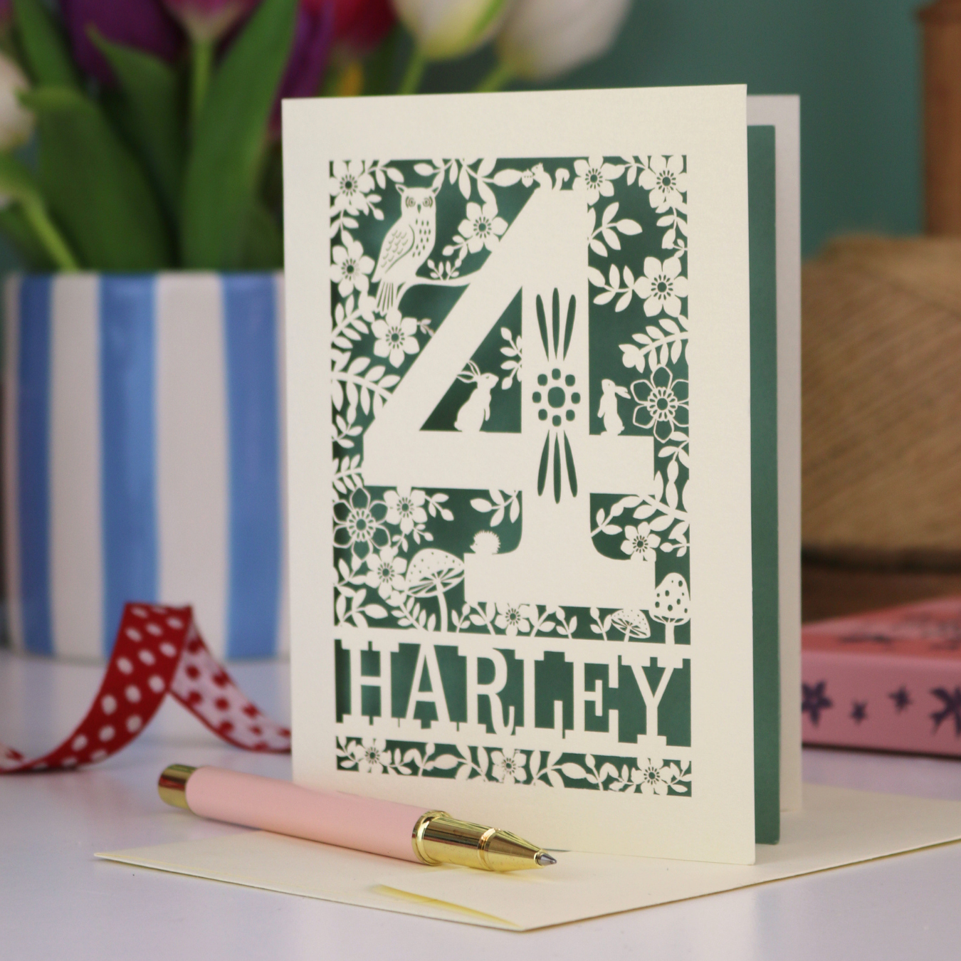 Decorative 4th Birthday card with woodland laser cut patterns, personalised with the name 'Harley', on a table with stationery items.