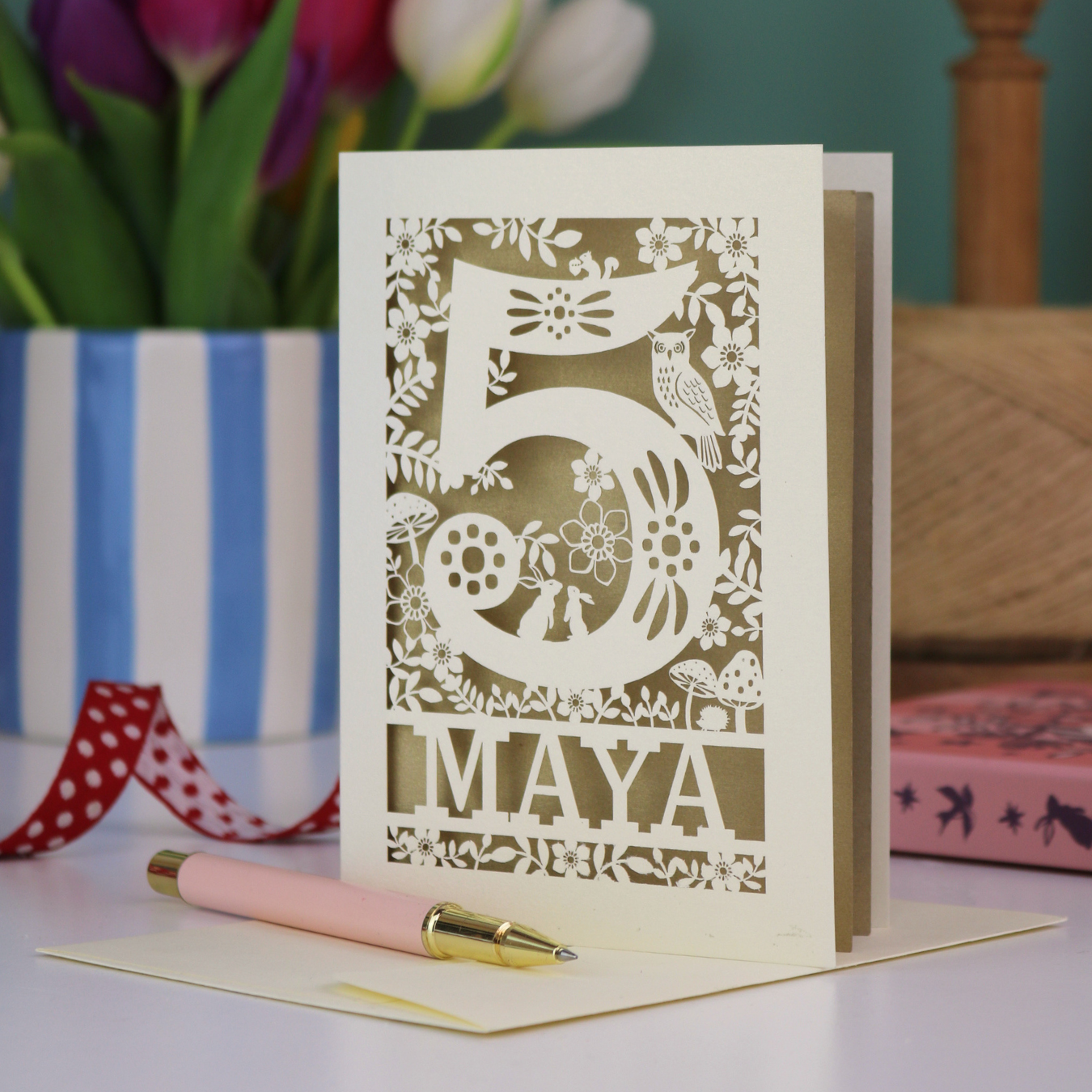 Decorative child's 5th birthday card with '5 MAYA' and woodland laser cut design on a table with flowers and books in the background