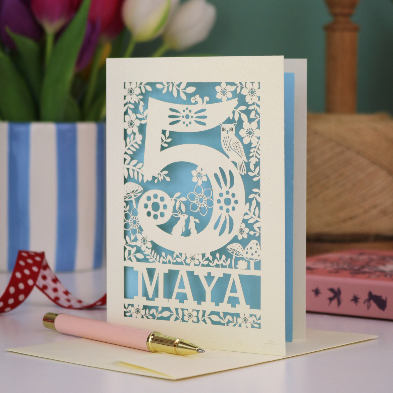 Decorative child's 5th birthday card with '5 MAYA' and woodland laser cut design on a table with flowers and books in the background
