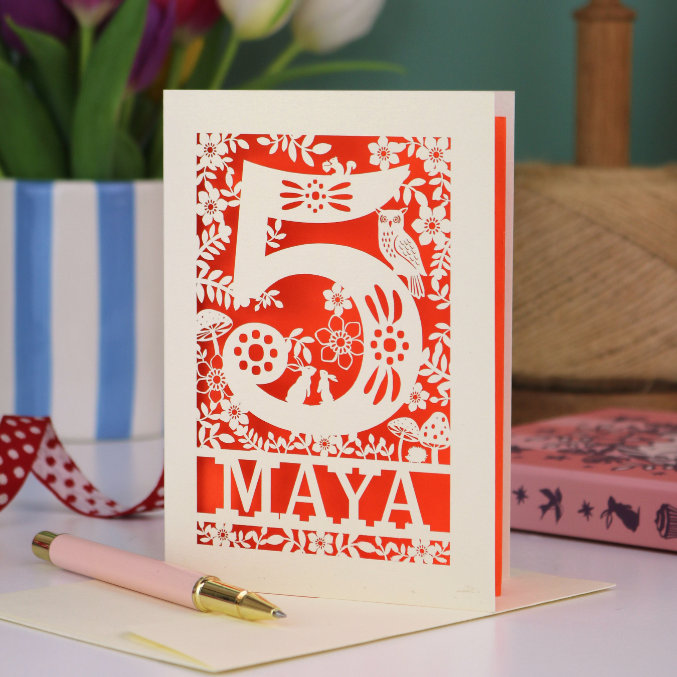 Decorative child's 5th birthday card with '5 MAYA' and woodland laser cut design on a table with flowers and books in the background