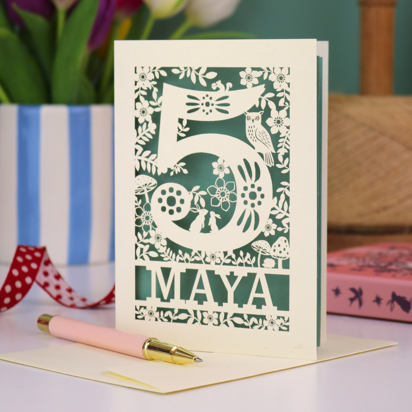 Decorative child's 5th birthday card with '5 MAYA' and woodland laser cut design on a table with flowers and books in the background