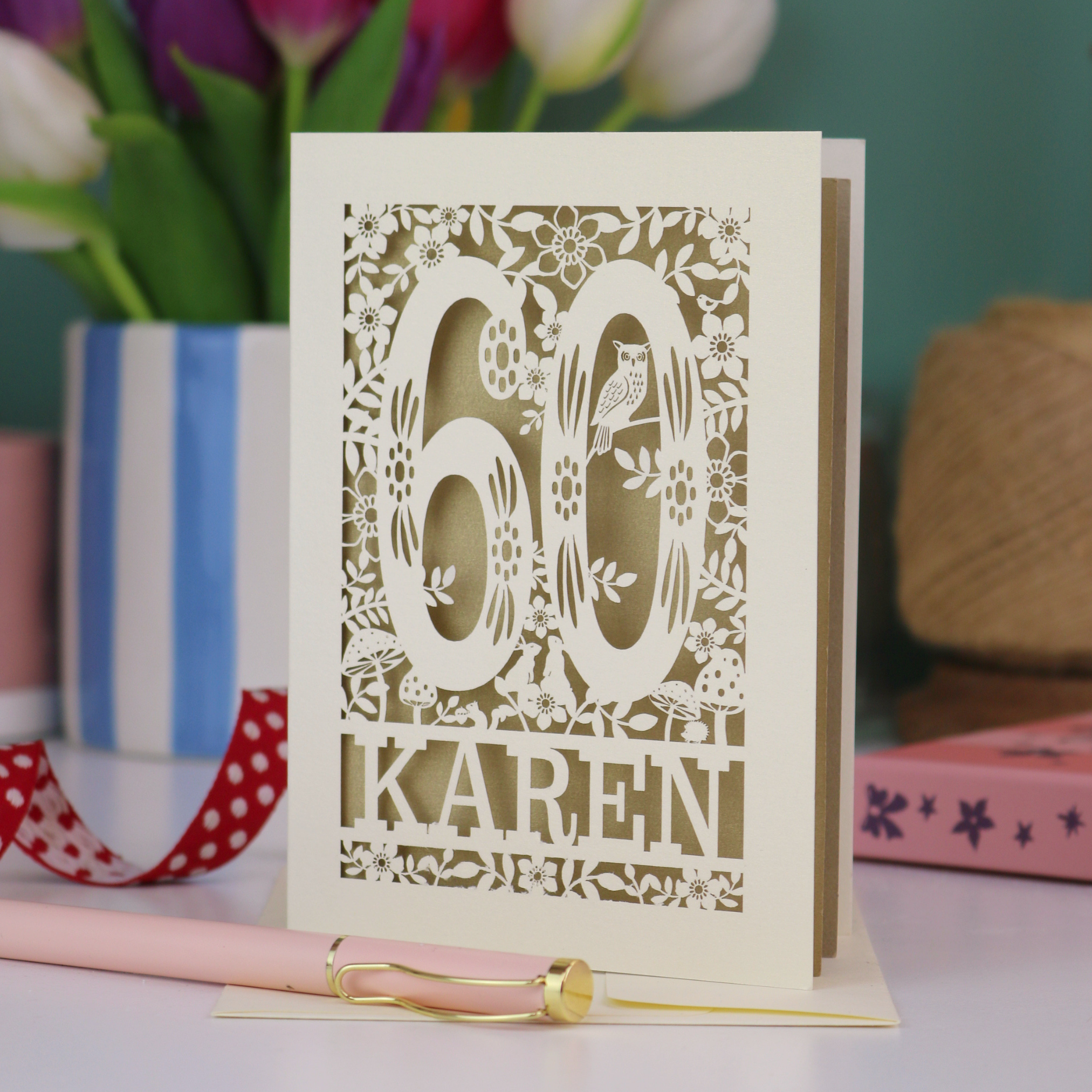 A laser cut 60th birthday card with woodland design and decorative number '60', personalised with the name "Karen". The birthday card is laser cut from a metallic cream card and has a gold leaf coloured paper insert. It stands on a white desk with flowers and stationery items. 