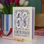 A laser cut 60th birthday card with woodland design and decorative number '60', personalised with the name "Karen". The birthday card is laser cut from a metallic cream card and has a lilac paper insert. It stands on a white desk with flowers and stationery items. 
