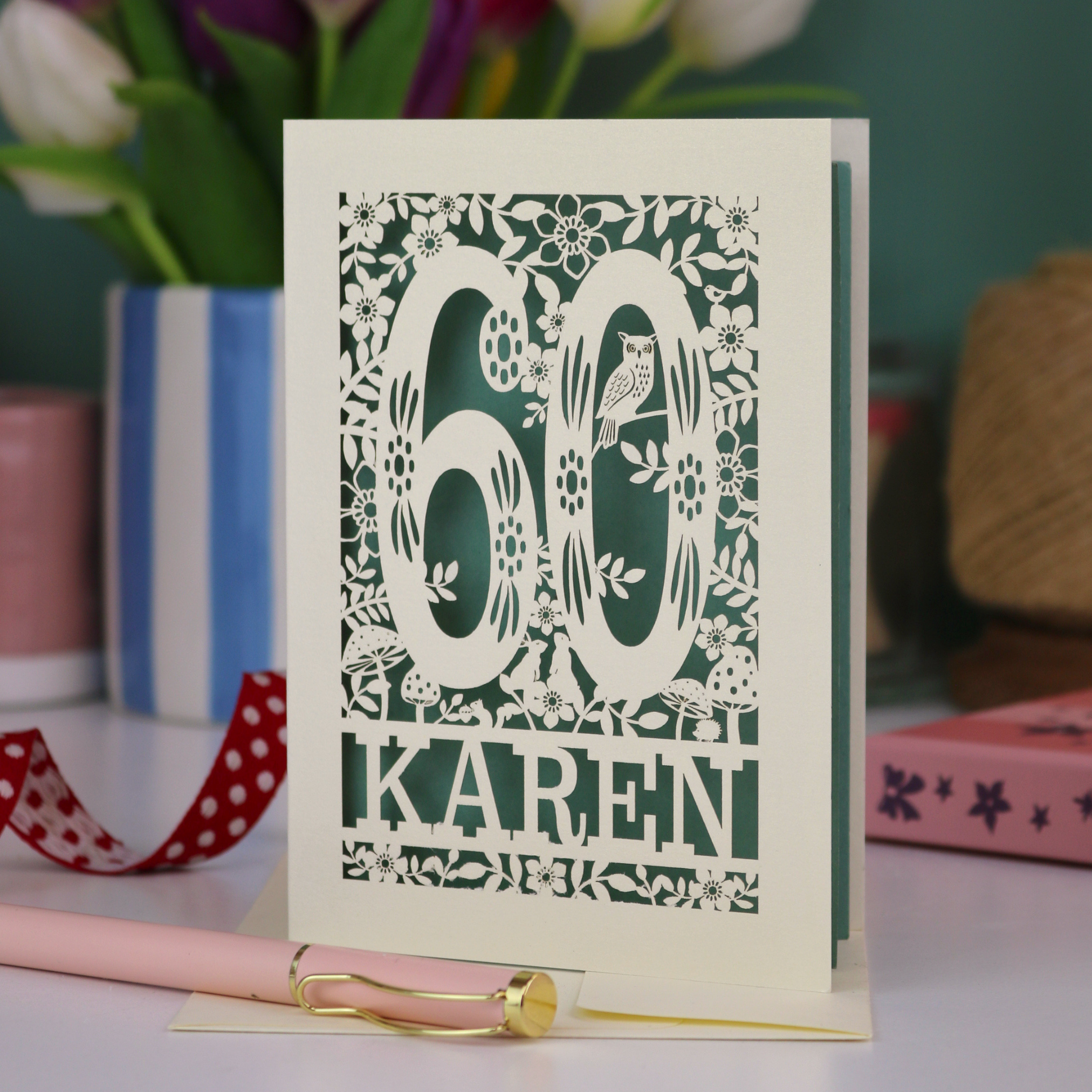 A laser cut 60th birthday card with woodland design and decorative number '60', personalised with the name "Karen". The birthday card is laser cut from a metallic cream card and has a sage paper insert. It stands on a white desk with flowers and stationery items. 