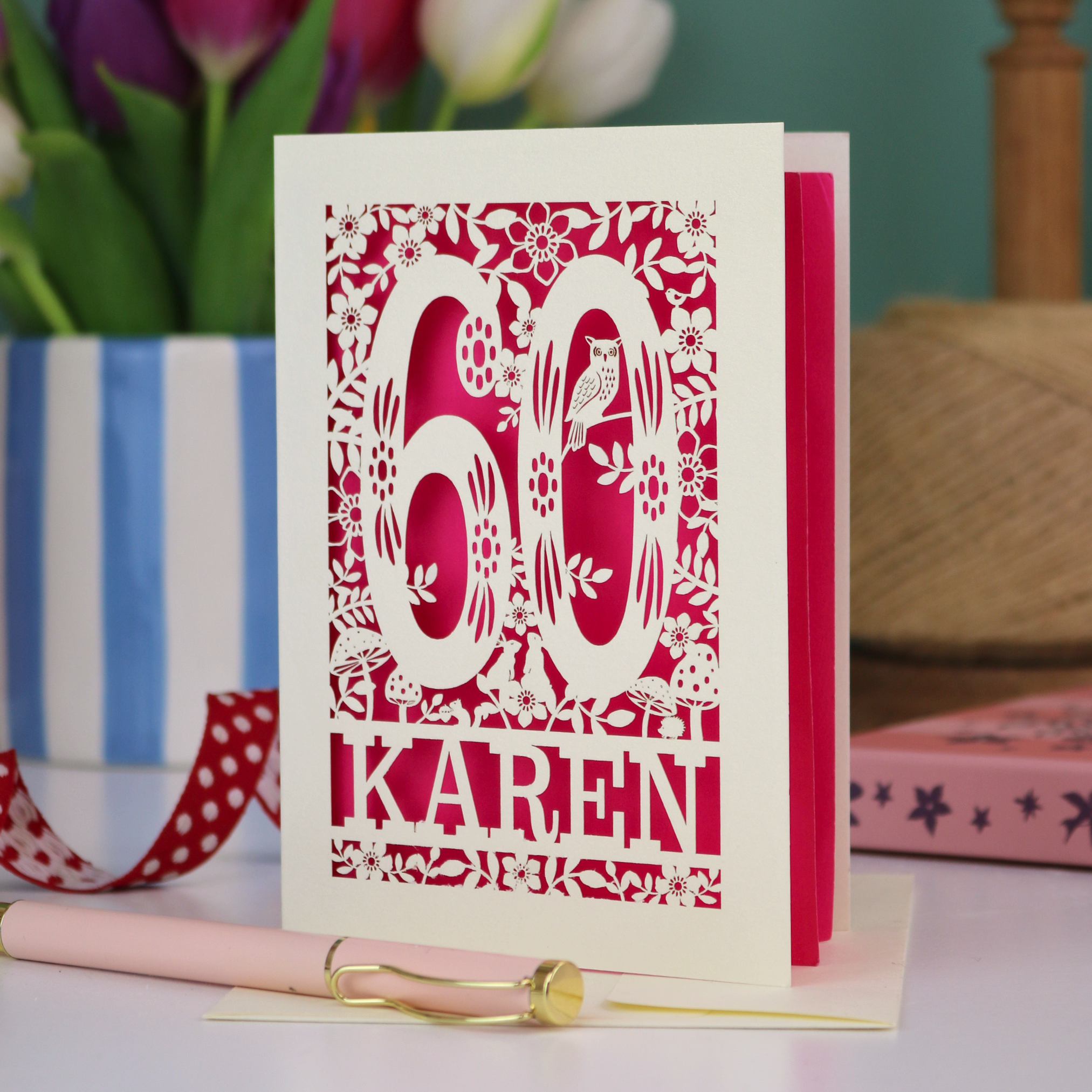 A laser cut 60th birthday card with woodland design and decorative number '60', personalised with the name "Karen". The birthday card is laser cut from a metallic cream card and has a shocking pink paper insert. It stands on a white desk with flowers and stationery items. 