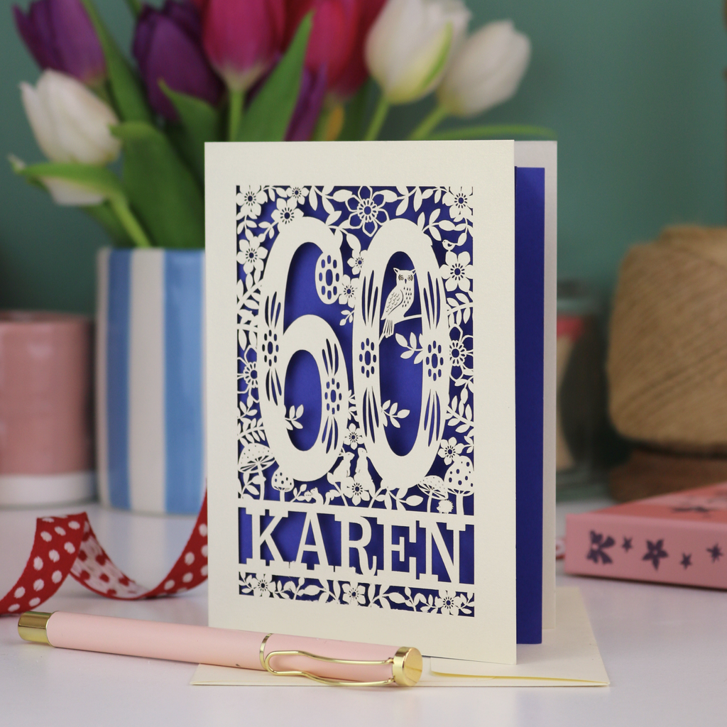 A laser cut 60th birthday card with woodland design and decorative number '60', personalised with the name "Karen". The birthday card is laser cut from a metallic cream card and has a violet paper insert. It stands on a white desk with flowers and stationery items. 
