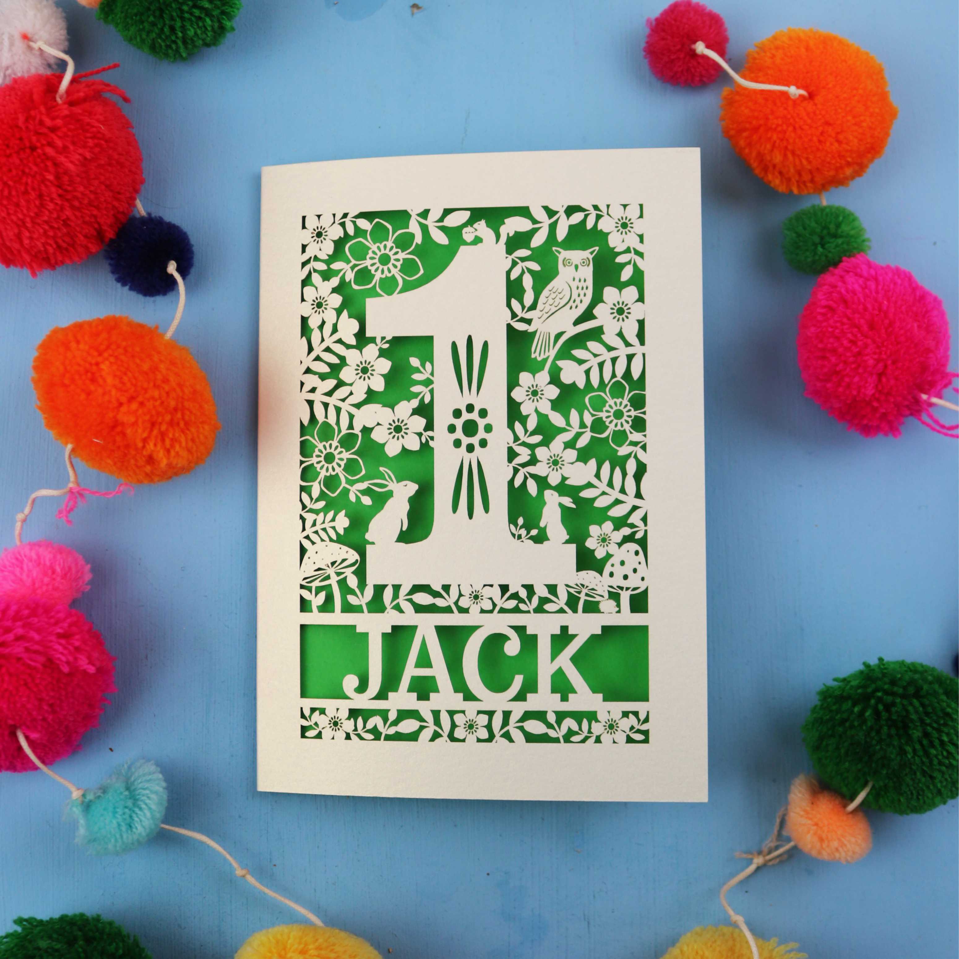 Personalised Laser cut First Birthday Card with 'Jack' design surrounded by colourful pom-poms on a blue background