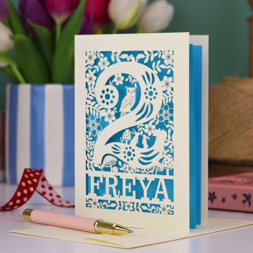 A laser cut 2nd birthday card with woodland design and decorative '2', personalised with a name. The card has tiny cut out woodland elements including bunnies, toadstools, squirrel and owl.
