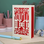Laser cut funny valentine's Card with 'YOU'LL DO' text and floral patterns on a desk with stationery items.
