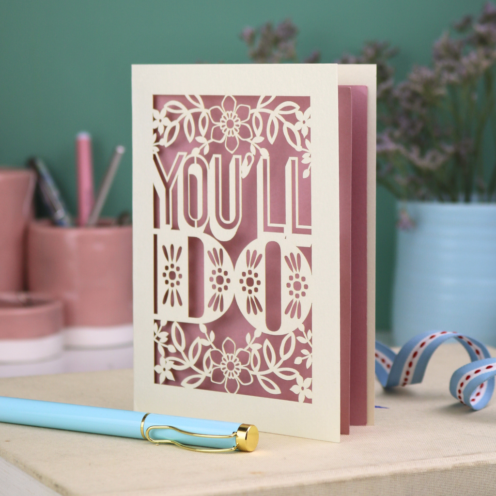Cut out funny valentine's Card with 'YOU'LL DO' text and floral patterns on a desk with stationery items.