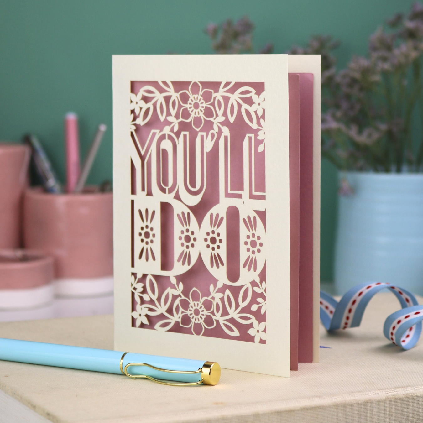 Cut out funny valentine's Card with 'YOU'LL DO' text and floral patterns on a desk with stationery items.