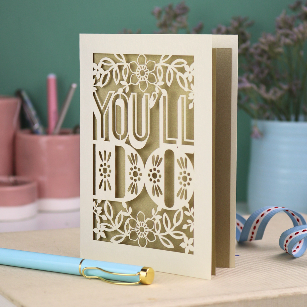 Laser cut funny valentine's Card with 'YOU'LL DO' text and floral patterns on a desk with stationery items.