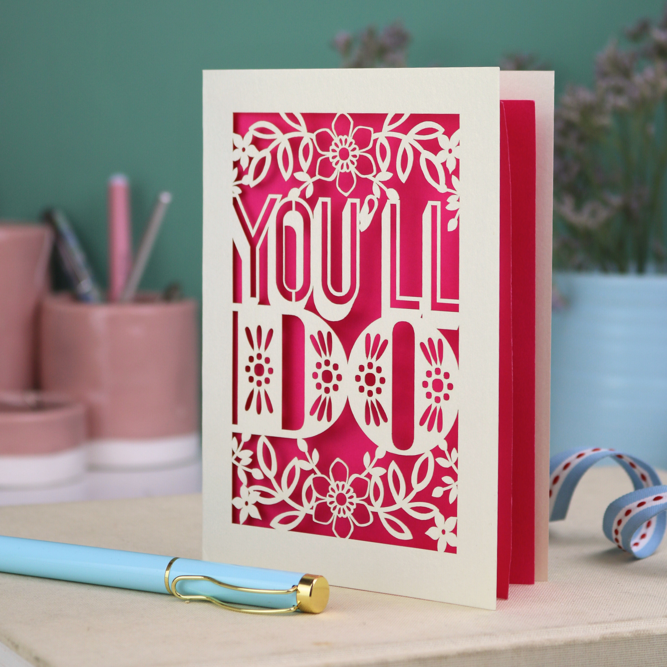 Laser cut funny valentine's Card with 'YOU'LL DO' text and floral patterns on a desk with stationery items.
