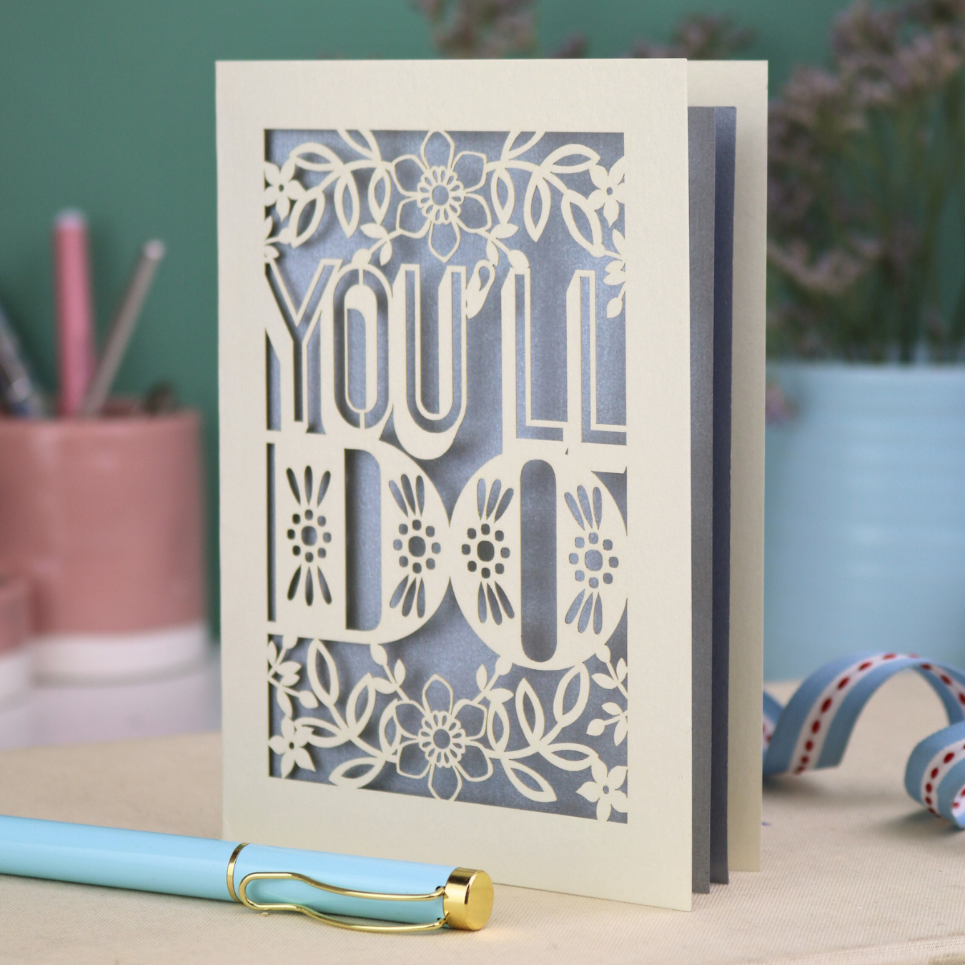 Paper cut funny valentine's Card with 'YOU'LL DO' text and floral patterns on a desk with stationery items.
