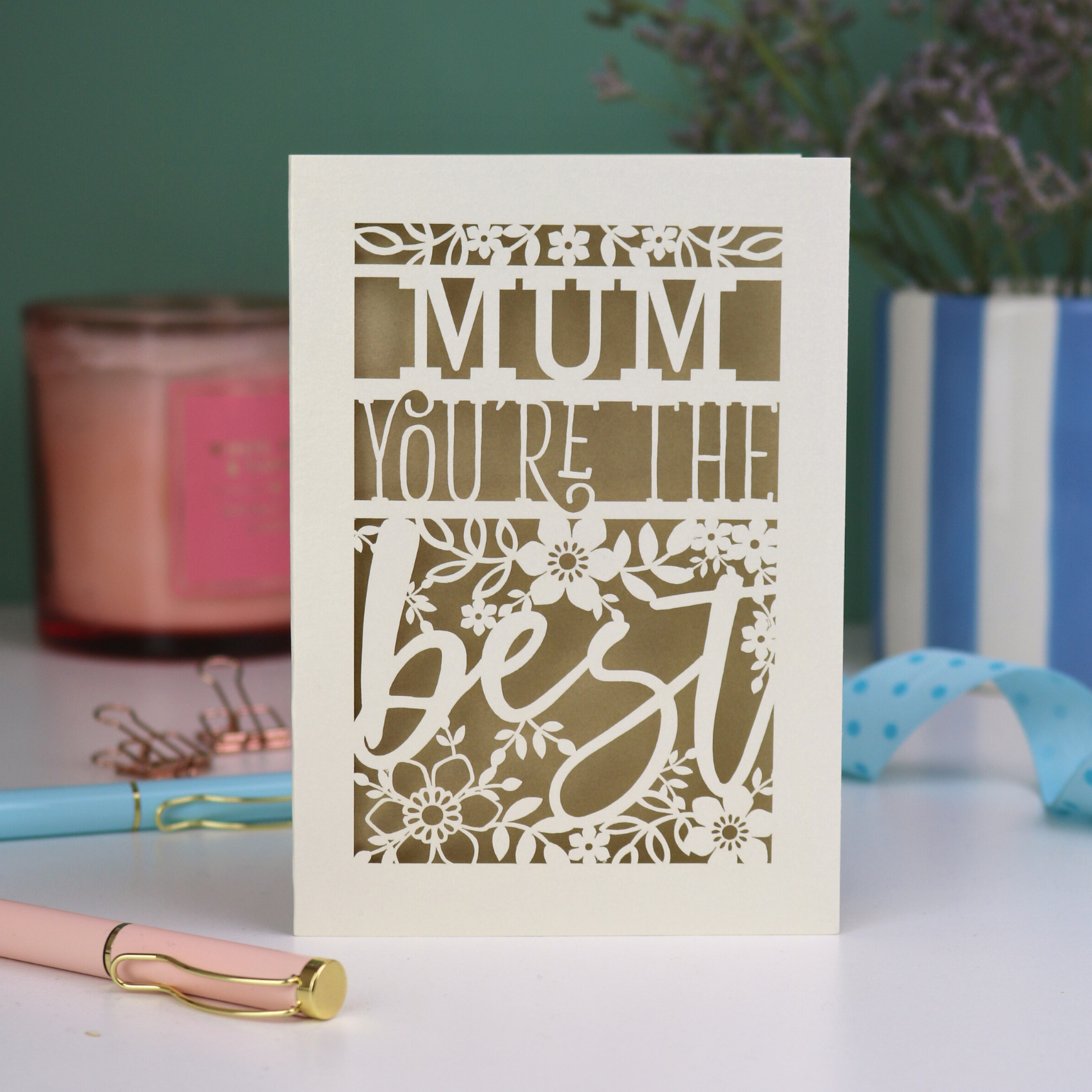 A laser cut Mother's Day card that says 'Mum, You're the best', with floral design elements. Card stands on a white desk with stationery items