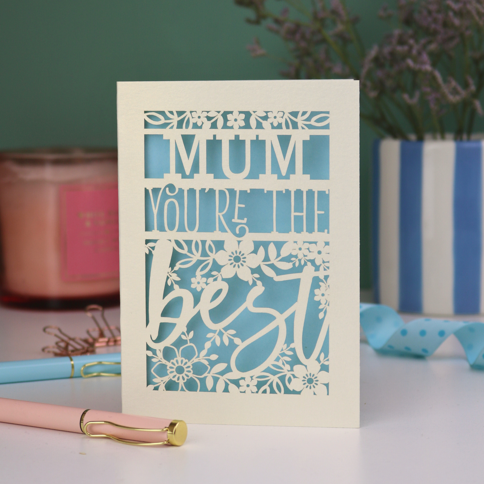 A personalised laser cut Mother's Day card that says 'Mum, You're the best', with floral design elements. Card stands on a white desk with stationery items