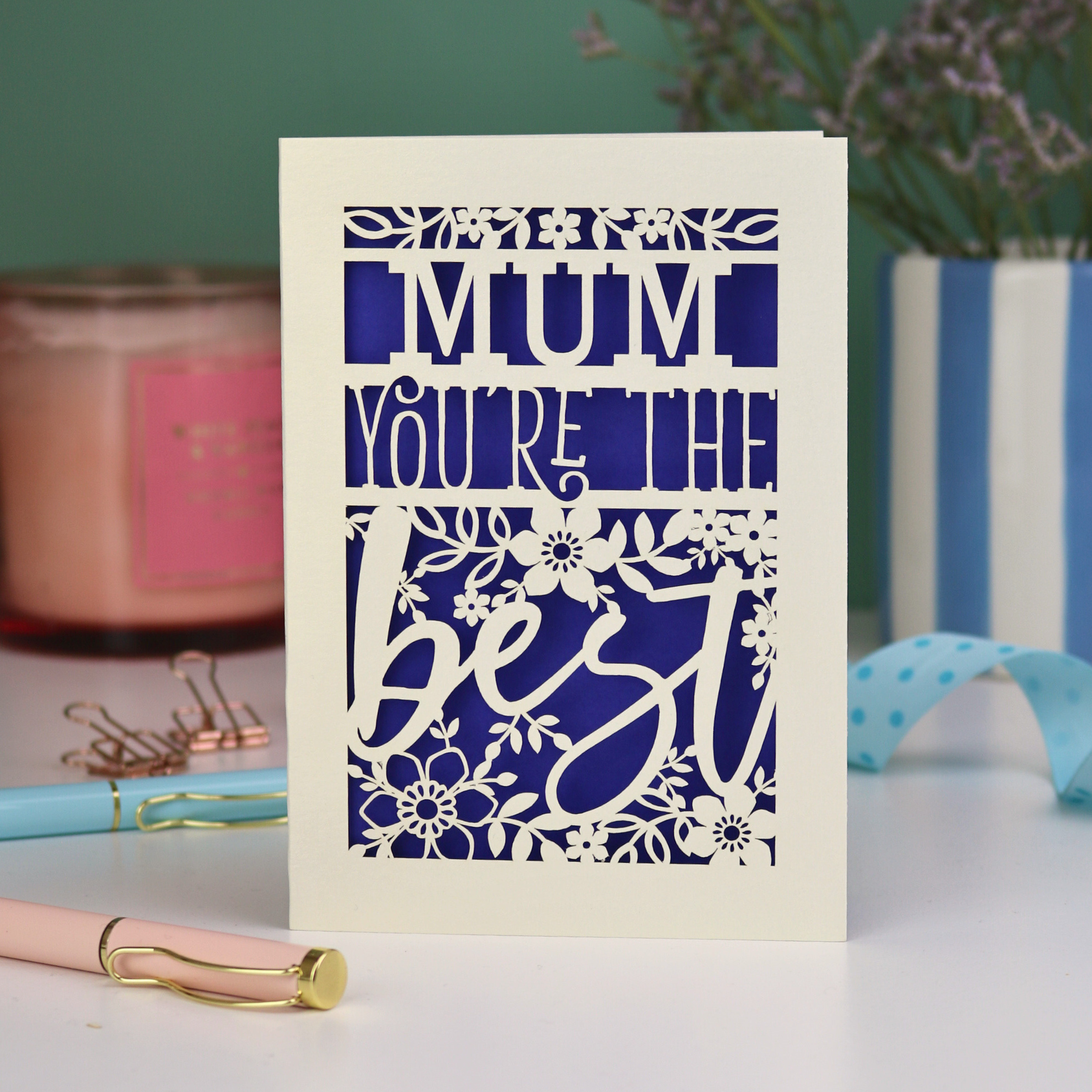A laser cut Mother's Day card that says 'Mum, You're the best', with floral design elements. Card stands on a white desk with stationery items