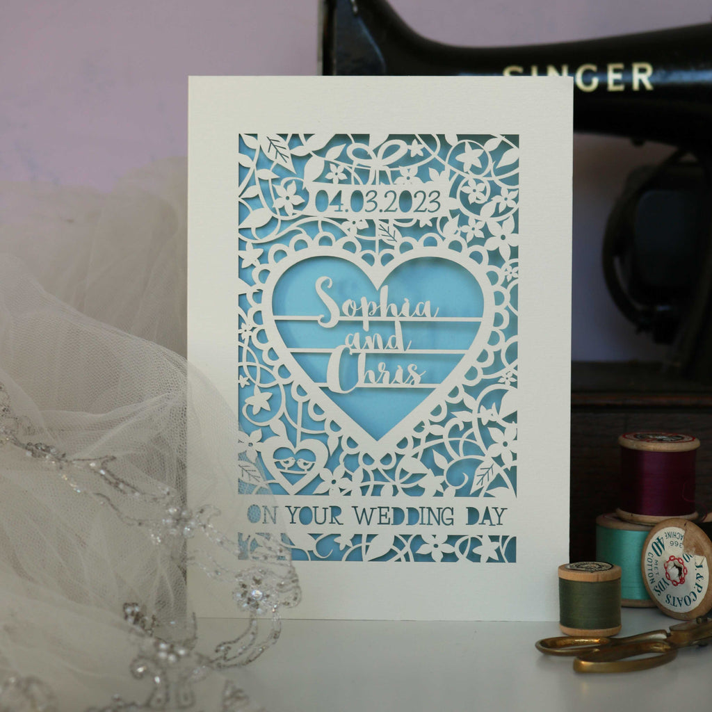 A wedding card that is laser cut and personalised and will make the couple cry. Seriously, it's the most beautiful card. It's laser cut from cream card and has a light blue paper insert. The design is a flowery leafy border with a big heart in the centre. In the heart are 3 lines of text with the names "Sophia & Chris". There is a space for the date of the wedding and the card says "On your wedding day" at the bottom in cut out writing.