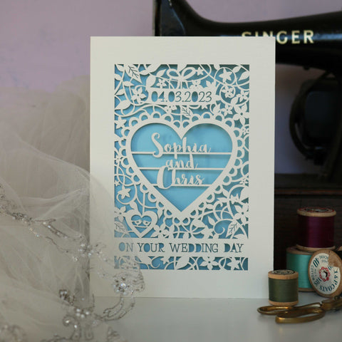 A wedding card that is laser cut and personalised and will make the couple cry. Seriously, it's the most beautiful card. It's laser cut from cream card and has a light blue paper insert. The design is a flowery leafy border with a big heart in the centre. In the heart are 3 lines of text with the names "Sophia & Chris". There is a space for the date of the wedding and the card says "On your wedding day" at the bottom in cut out writing. - A6 (small) / Light Blue