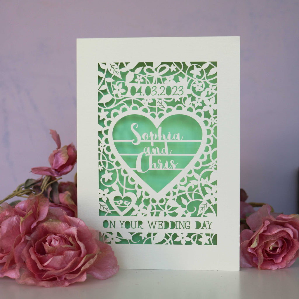 Laser cut wedding cards, cut from cream card with a light green paper insert behind.  Card features floral shapes around a heart with the names "Sophia & Chris". The date is in a banner at the top, and a line of text at the bottom reads "On Your Wedding Day."