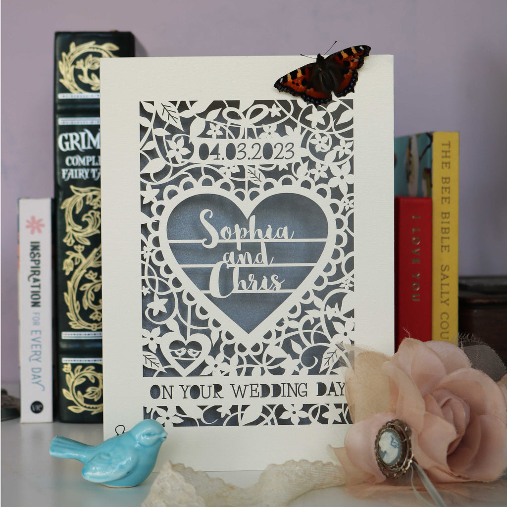 A personalised papercut wedding card featuring the date in numbers, two names and the words "On Your Wedding Day" surrounded by flowers, leaves and two tiny birds inside a heart.