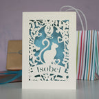 Papercut birthday card showing a cat surrounded with flowers and a balloon. Cut from cream card and finished with pale blue paper. Personalised with a name and age.