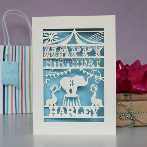 Big Top themed personalised papercut birthday card.  Shows an elephant and seals under the big top. Shown  laser cut from cream card with a pale blue background and personalised with age and name. - A6 (small) / Light Blue