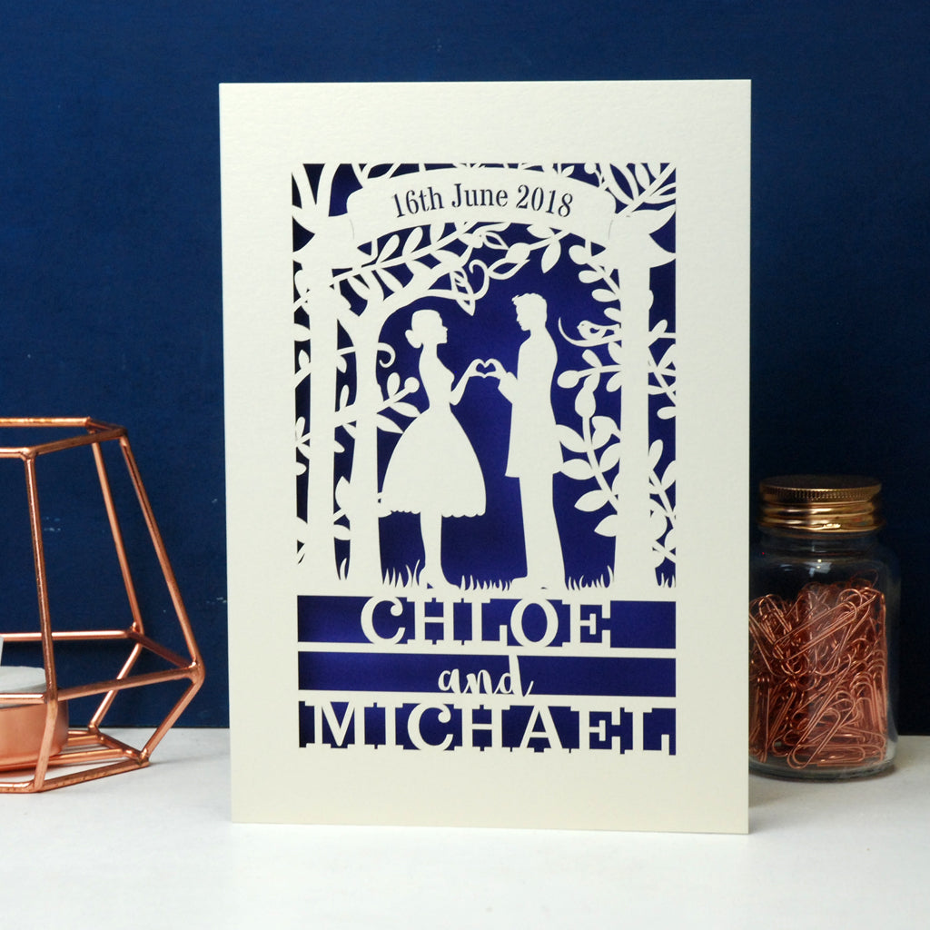 A laser cut engagement card that features the silhouettes of a couple, and is personalised with a date in a banner at the top and the names of the couple over three lines of text at the bottom