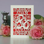 A cream laser cut engagement card with a red paper insert. Card has two names and a large, floral ampersand, with flowers and leaves around it.