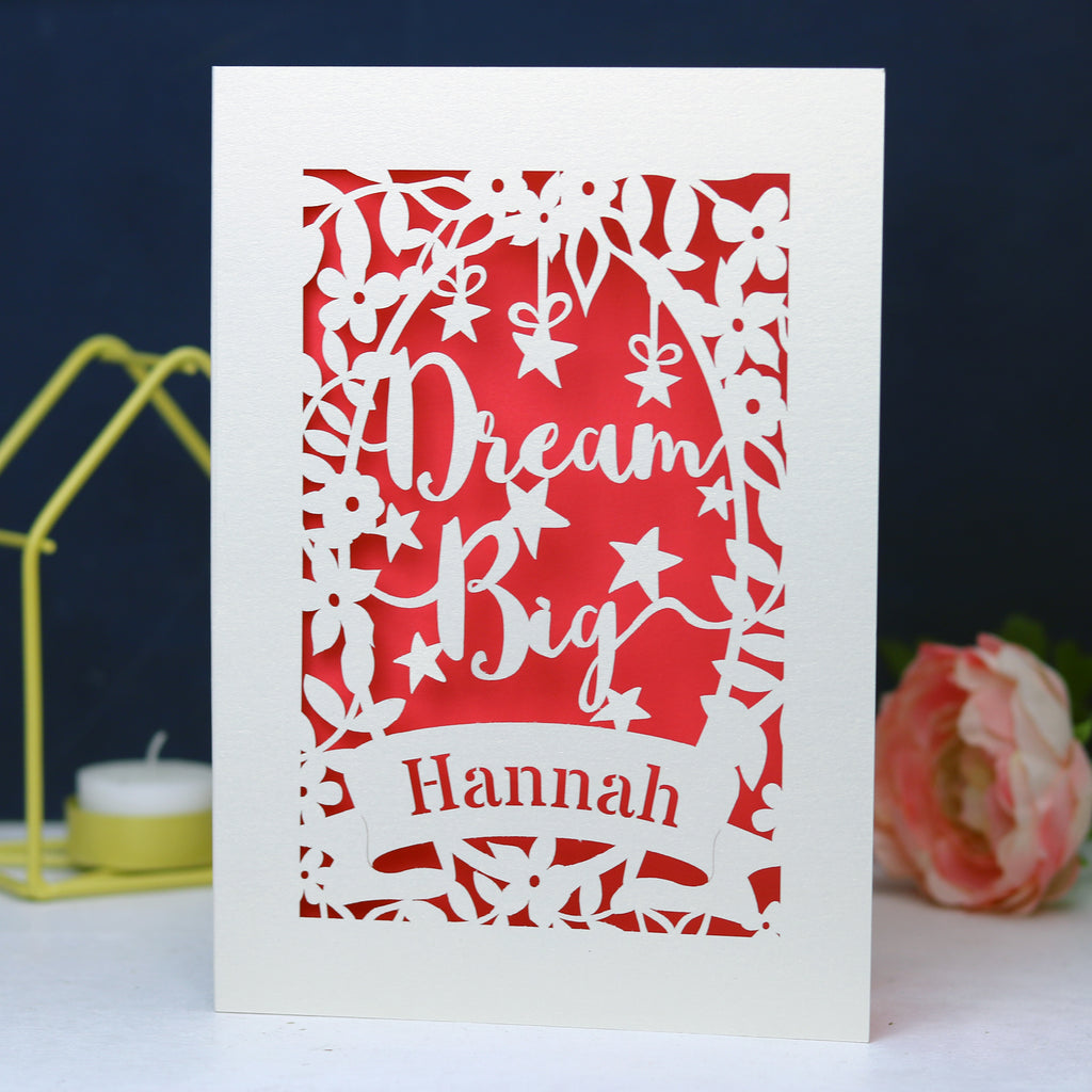A personalised laser cut card with the words 'Dream Big' and a name 'Hannah' written on it, displayed on a dark background surrounded by colourful pompons.