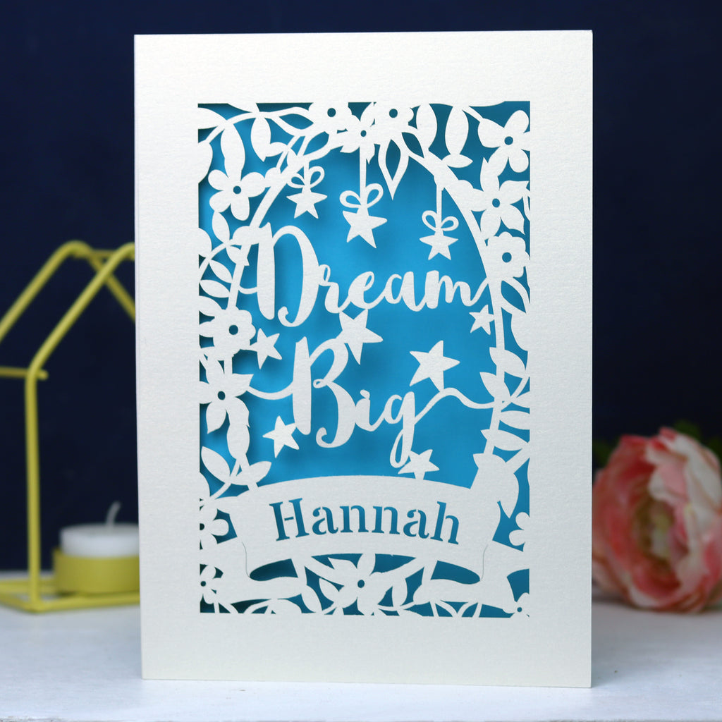 A personalised laser cut card with the words 'Dream Big' and a name 'Hannah' written on it, displayed on a dark background surrounded by colourful pompons.