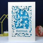 A personalised laser cut card with the words 'Dream Big' and a name 'Hannah' written on it, displayed on a dark background surrounded by colourful pompons.