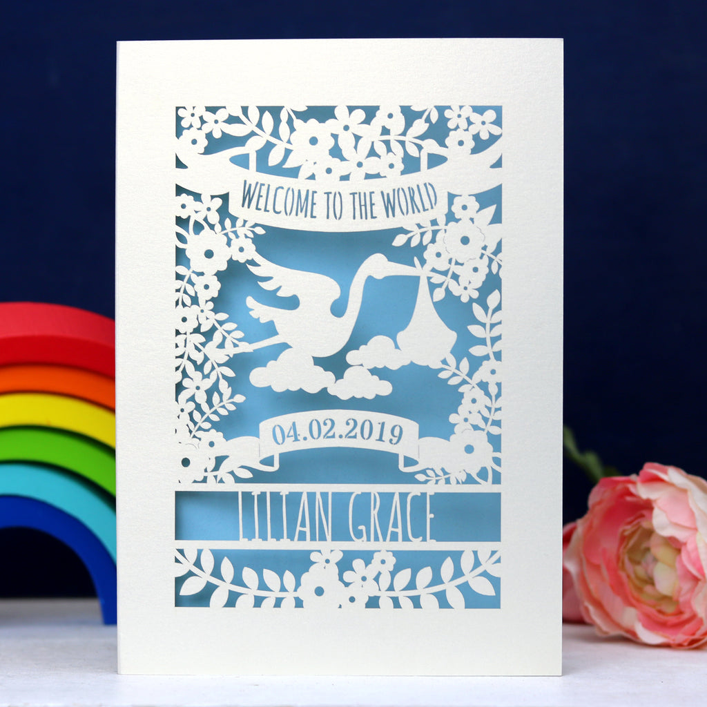 A cream and light blue papercut baby card personalised with a name and date. Top of the card reads "Welcome to the World" in a banner. Underneath the banner is a stork, clouds, leaves and flowers. 
