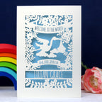 A cream and light blue papercut baby card personalised with a name and date. Top of the card reads "Welcome to the World" in a banner. Underneath the banner is a stork, clouds, leaves and flowers. 