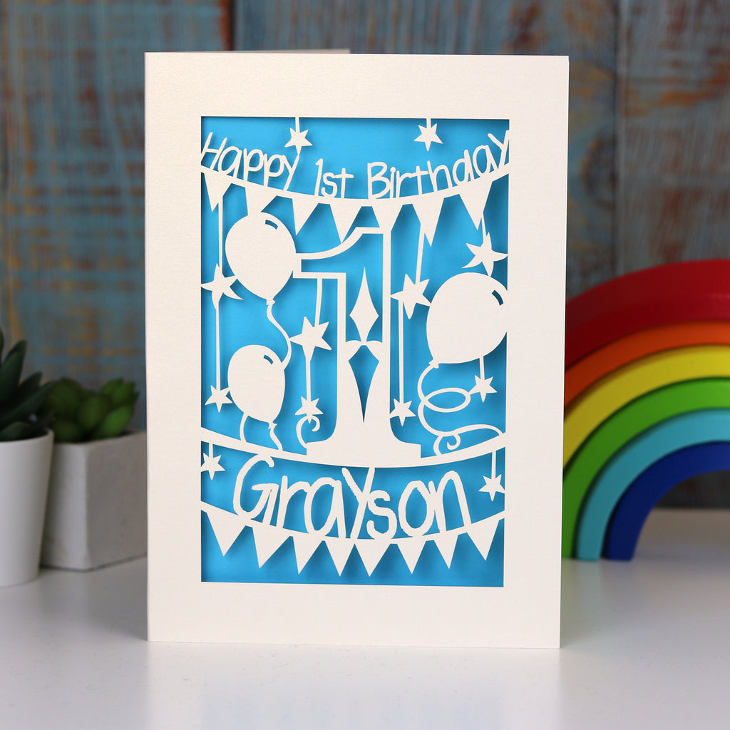 Personalised first birthday paper cut card, cream and peacock blue. Card is laser cut and reads Happy Birthday with a name underneath.