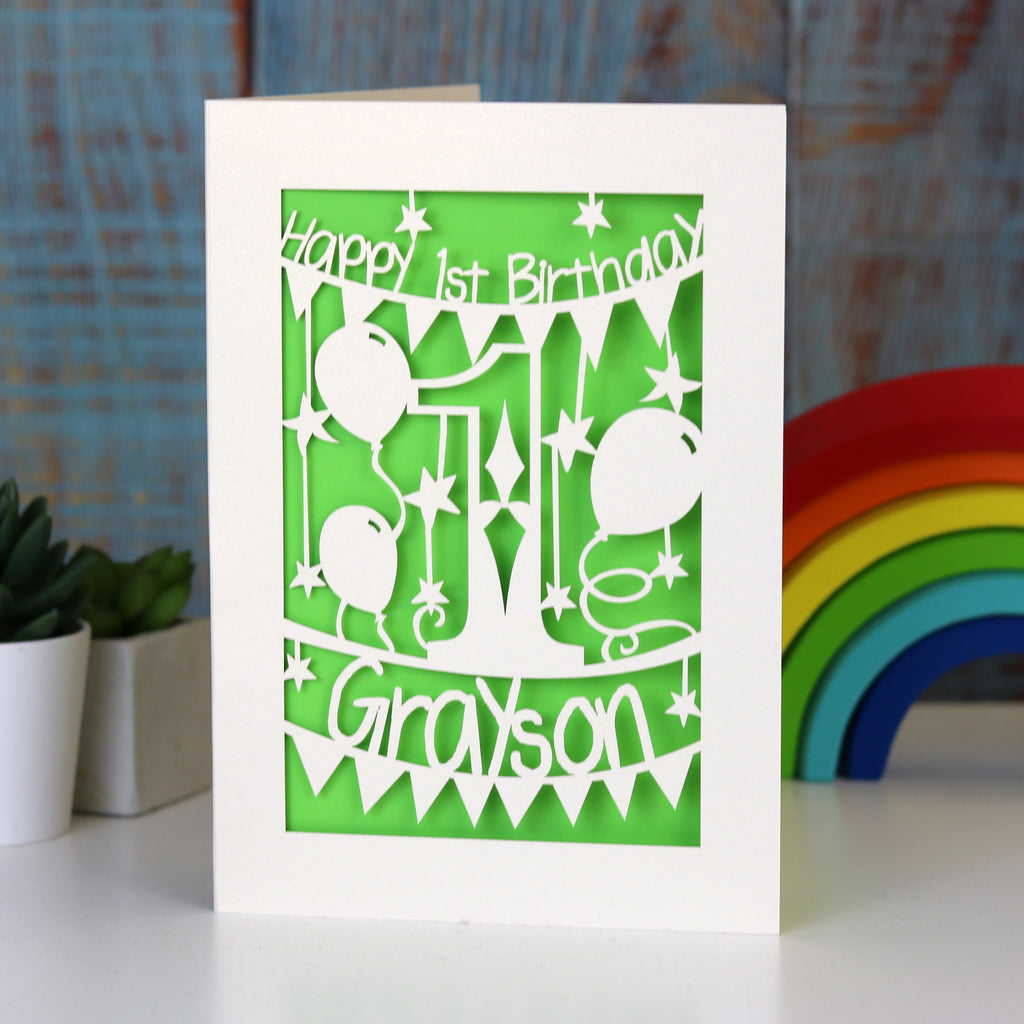 A cream and green paper cut first birthday card for grandson or granddaughter. Card is personalised with a name.