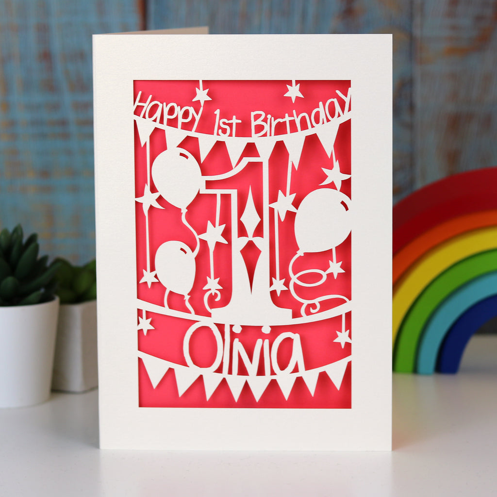 A coral pink 1st birthday card for grandchild. Happy 1st Birthday is written at the top and a name is at the bottom.