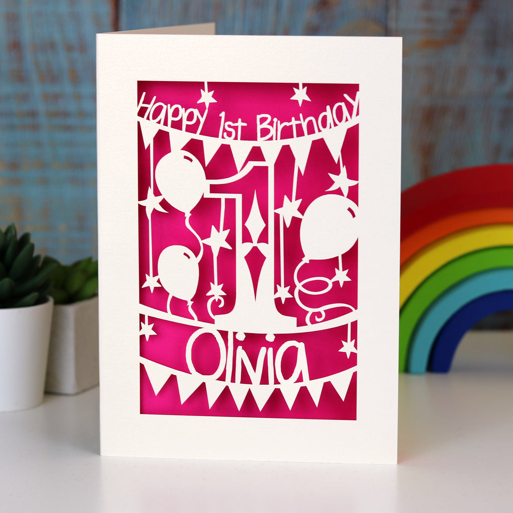 A laser cut card with a number 1 in the middle. Cut out card reveals a shocking pink paper behind. Card says Happy first Birthday with a name.