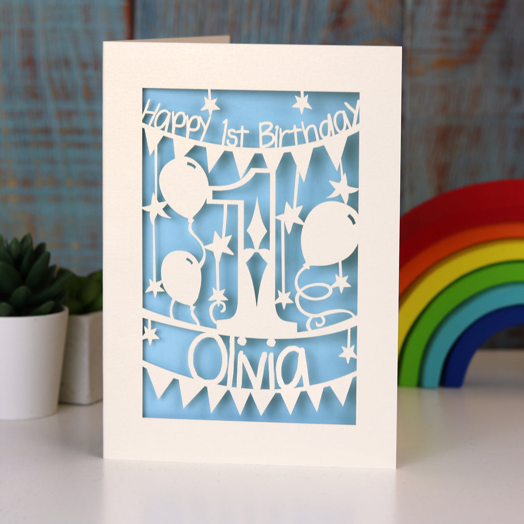 A laser cut first birthday with a cream cut card design and light blue paper insert. Card says "Happy 1st Birthday" and features a number 1 and balloons, with the child's name underneath. 