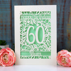 A laser cut cream flowery birthday card with happy birthday, 60, mum on it. The card has a light green paper insert behind the cut card