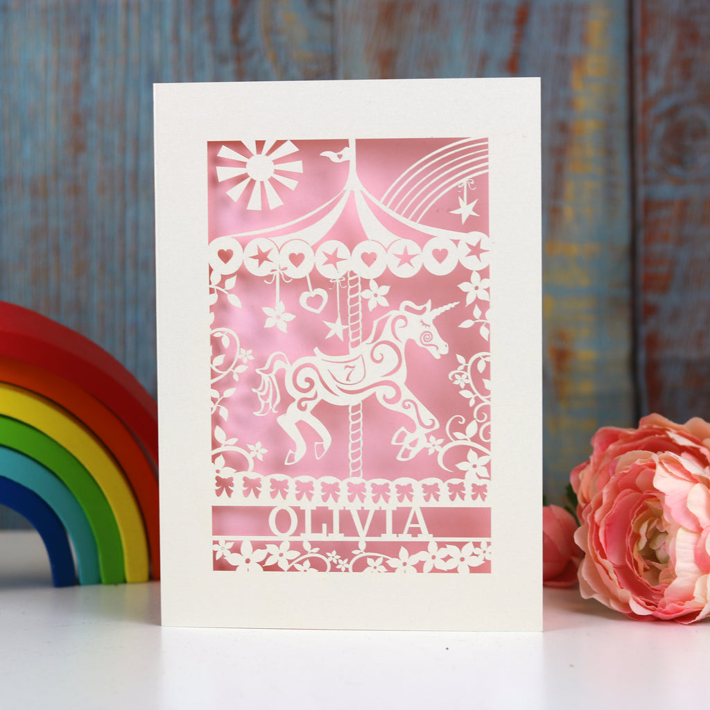A cream and light pink girls birthday card with a carousel horse, a name and an age.