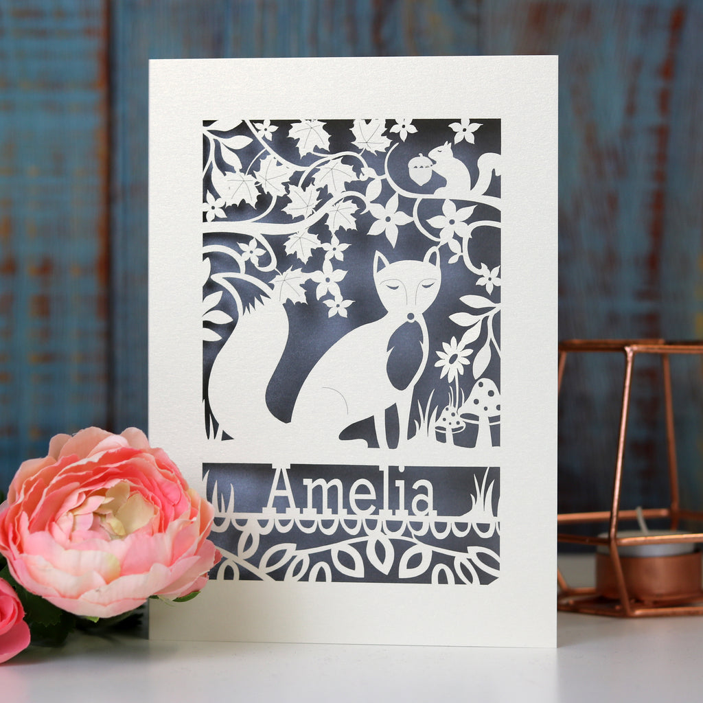 A fox birthday card, laser cut and with a silver paper insert. There is a fox in the centre, flowers and leaves around, with a squirrel and toadstool details. The name is under the fox.