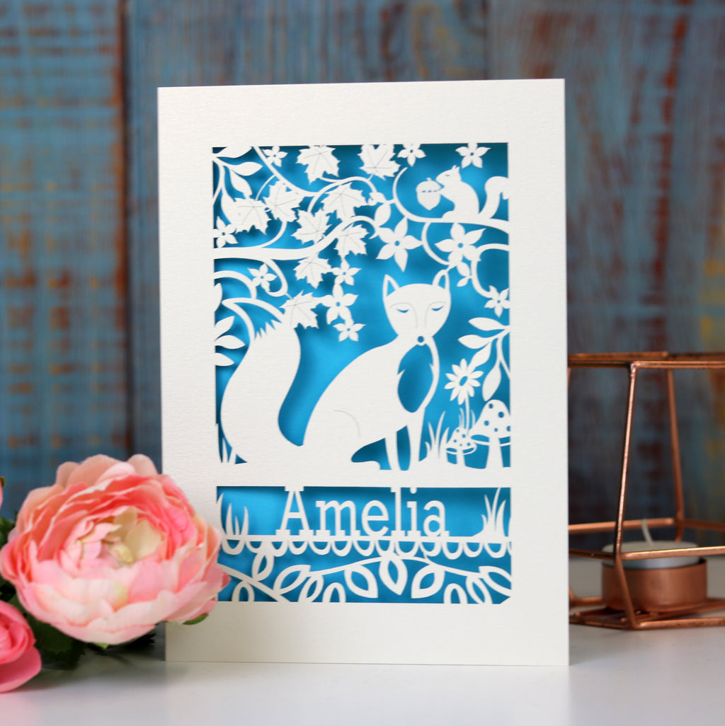 A laser cut card for birthdays. This card has a fox and a name and flower and leaf shapes. Card is cut away to reveal a blue paper behind