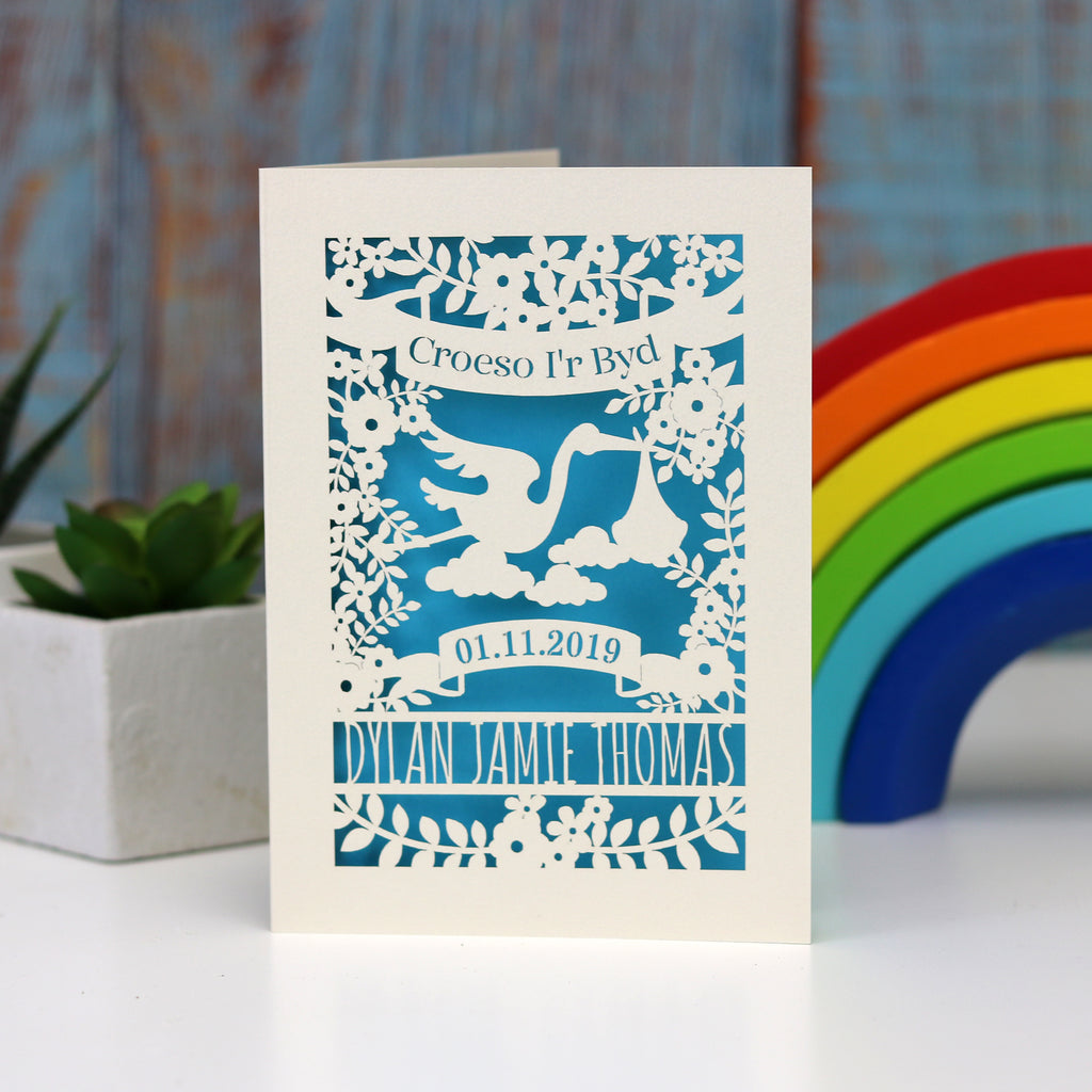 Laser cut personalised new baby card in Welsh. Shows  Croeso i'r Byd in a banner above a stork carrying a baby bundle. Lines below for the baby's name and a banner for the date of birth. Shown in cream with peacock blue insert paper.