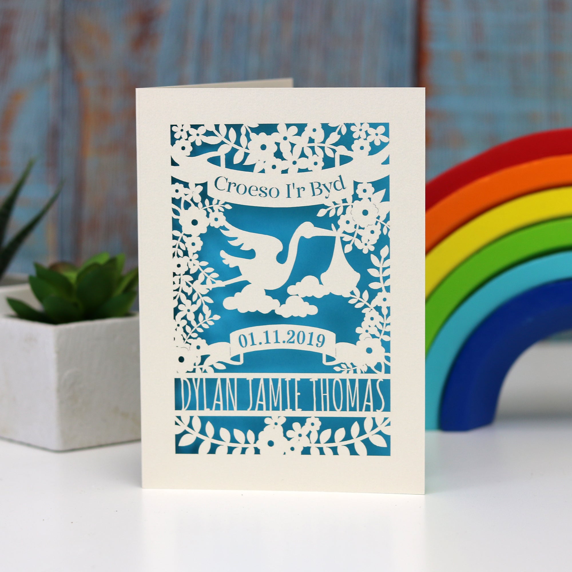 Laser cut personalised new baby card in Welsh. Shows  Croeso i'r Byd in a banner above a stork carrying a baby bundle. Lines below for the baby's name and a banner for the date of birth. Shown in cream with peacock blue insert paper.