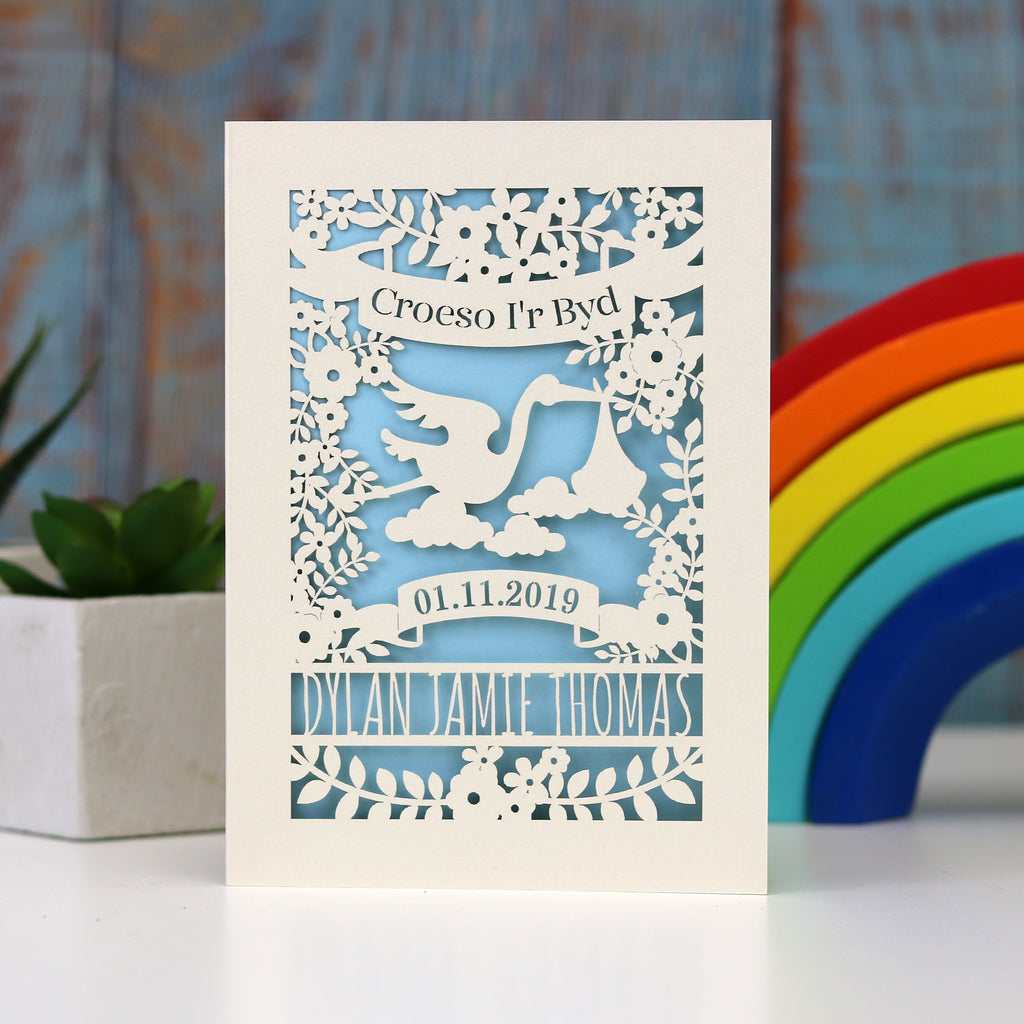 Personalised  papercut new baby showing Welcome to the World in Welsh, Croeso i'r Byd, especially for Welsh new babies!  Laser cut from cream card and finished with a baby blue insert. Shows a stork, flowers, leaves and space for the baby's name and date of birth.