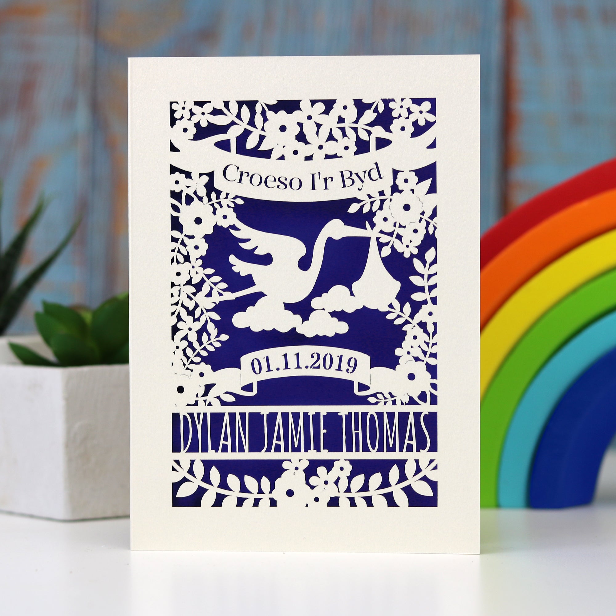 Croeso i'r Byd, welcome to the world in Welsh, personalised papercut new baby card especially for Welsh customers!  This card shows a stork flying with a baby  bundle, surrounded with flowers and space for baby's name and date of birth.  Cut from cream card, with a dark blue insert paper.